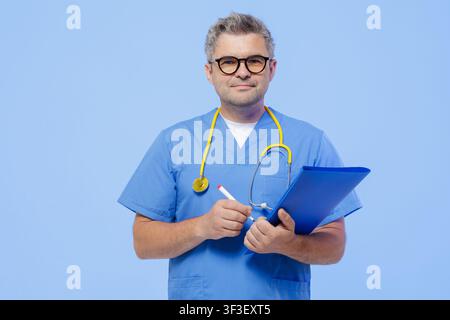 A professional doctor in blue scrubs, holding a clipboard and pen, ready for a patient's appointment in a modern medical setting. Stock Photo