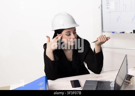 Female engineer in white hardhat sitting at desk touching head confused Stock Photo
