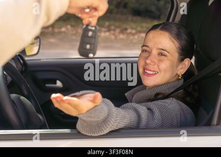 Smiling young woman looking at car key being handed to her through window, car interior, achievement concept Stock Photo
