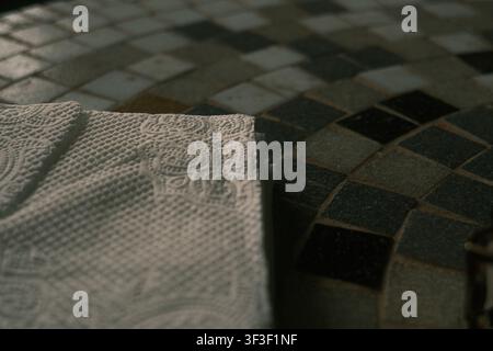 Close-up macro shot of textured, embossed white paper napkins resting on a dark geometric mosaic tile table under soft cinematic lighting. Stock Photo
