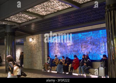 The interior of an upscale public transit space, likely Tokyo Station in Japan, featuring people seated on a stone bench beneath a large illuminated Stock Photo