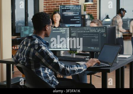 Programmer fixes his glasses while typing on a laptop and checking compiled code results at a desk with multiple software screens. Stock Photo