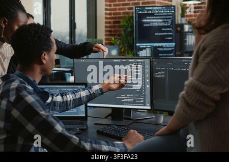 Software developers review source code compiling errors with the team, asking for explanations in front of multiple screens running algorithms. Stock Photo