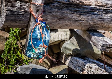 A blue plastic bottle filled with lobster bands hangs from ropes attached to a weathered wooden post on Paddy Beach Road in Baxters Harbour, Nova Scotia, Canada. The makeshift structure, made from flotsam, stands at the edge of a stone wall near the shore of the Bay of Fundy Stock Photo