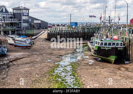 At low tide, the fishing boat ‘Custom Taylor’d II’ lies in the dry harbour basin at Halls Harbour, Nova Scotia, Canada. The boat’s name can be seen on the stern; the quay wall, made of wooden piles, withstands the tidal range of the Bay of Fundy, which is one of the largest in the world Stock Photo