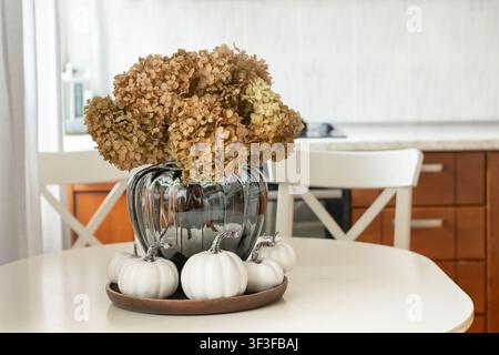 Elegant autumn home decor with dried hydrangea bouquet in glass vase and white decorative pumpkins on dining table in modern kitchen interior. Cozy se Stock Photo