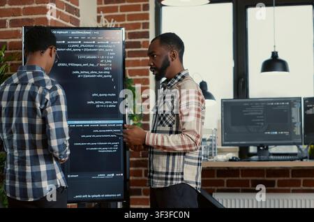 Software programmers leave after reviewing source code errors on a wall screen and digital tablet, showing teamwork in an artificial intelligence workflow. Stock Photo