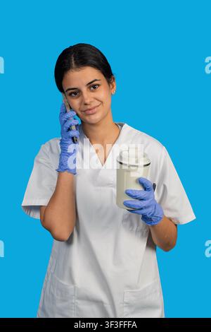 Young medical worker wearing gloves and uniform making a call on a blue background Stock Photo