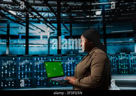 Engineer uses green screen laptop to implement security measures, fortifying data center defenses against cyber threats. Server hub worker uses chroma key notebook to detect threats, defending systems Stock Photo