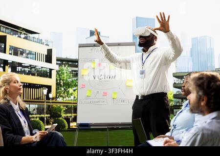 African american businessman wearing VR headset with hands raised, presenting strategy roadmap with interactive gadget and demonstration. Outdoor workshop beside whiteboard. Stock Photo