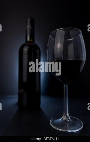 Moody close-up of a glass filled with red wine in sharp focus, with a wine bottle blurred behind it against a dark background and cool studio lighting Stock Photo