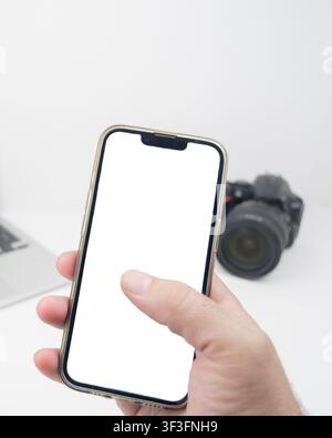 Hand using a smartphone with a blank white mockup screen, with a digital camera and laptop softly blurred in the background for mobile photography and Stock Photo