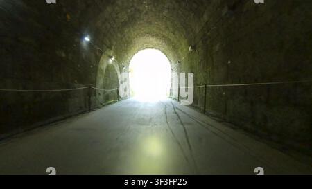A first person perspective captures the motion of riding through a dark, arched stone tunnel, moving toward a bright, glowing exit ahead. Stock Photo
