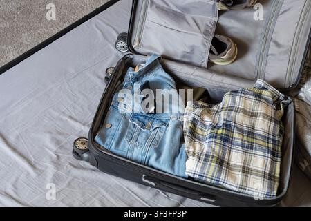 An open black suitcase lying on a grey bedsheet, carefully packed for a trip. Inside the luggage are a blue denim jacket, dark sunglasses, a plaid che Stock Photo
