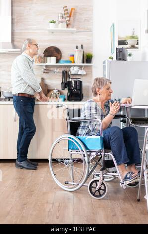 Disabled senior woman works from home on a laptop in the kitchen while her husband prepares breakfast, showing accessible remote work. Stock Photo