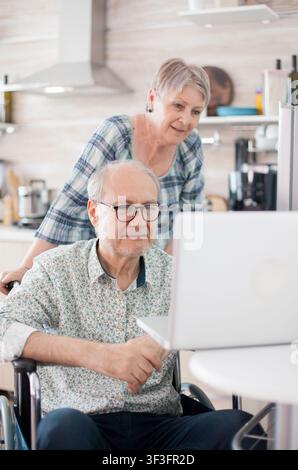 Disabled senior man in a wheelchair makes a family video call with his wife on a laptop in the kitchen, using online communication at home. Stock Photo