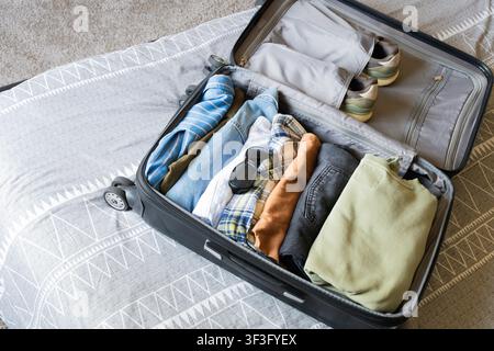 An open black suitcase lying on a grey duvet cover, carefully packed for a trip. Inside the luggage are shirts, jeans, blue denim jacket, sunglasses a Stock Photo