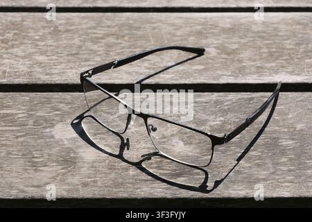 A pair of rectangular metal eyeglasses sits on a weathered wooden table, casting a distinct shadow in the sunlight Stock Photo