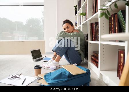Young woman student experiencing stress and anxiety while studying on the floor next to a bookshelf, She sits on the floor next to a bookshelf, surrou Stock Photo