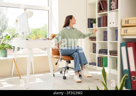 Young woman reaching for a book on a white bookshelf in a bright home office with a desk and computer, She is sitting at a modern white desk with a co Stock Photo