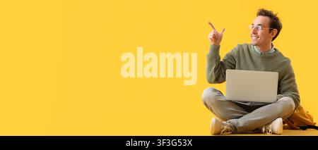 Smiling young man in glasses pointing up with a laptop, online education and remote work concept, yellow background Stock Photo