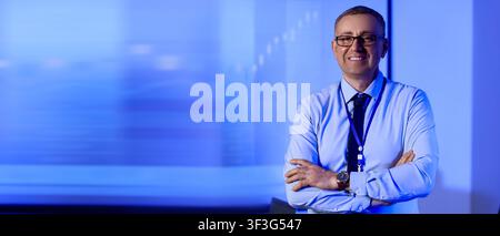 Smiling businessman in glasses and blue shirt with id badge, arms crossed, standing in a modern office with a blurred background Stock Photo