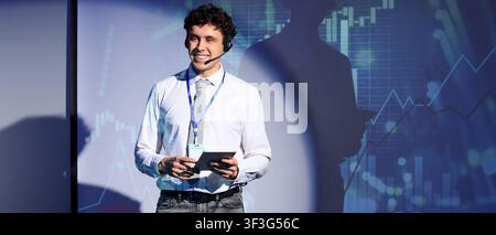 Smiling young male speaker with headset and tablet presenting financial data on a screen Stock Photo
