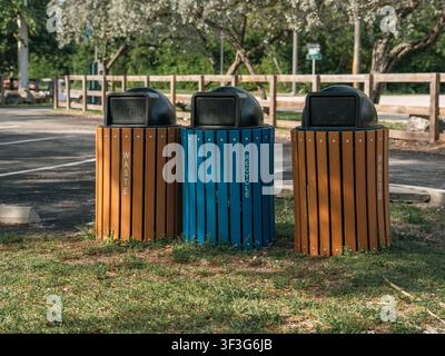 Three slatted outdoor disposal bins, including two brown waste receptacles and one blue recycling unit, stand side by side on a grassy median. Stock Photo