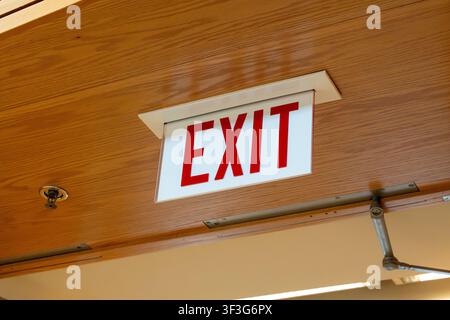 Illuminated red EXIT sign mounted on a wooden ceiling panel with door closer hardware, highlighting indoor safety systems, emergency guidance Stock Photo