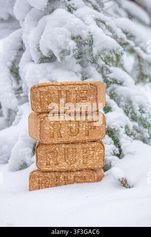 View of a stack of RUF wood fuel briquettes placed in deep snow with a snow-covered pine tree in the background in Latvia. Stock Photo
