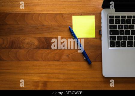 Laptop with a yellow sticky note and blue pen on a wooden desk, illustrating planning, productivity and office organization. Stock Photo