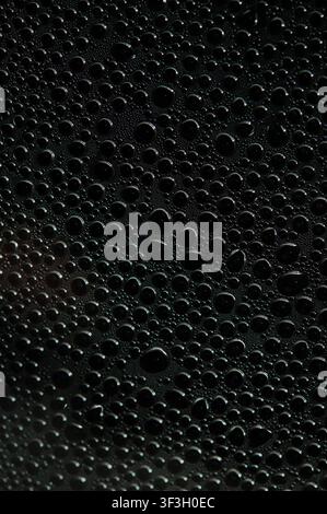 CAPTION (135 characters): Macro view of glossy black water droplets across a dark textured surface, forming an abstract pattern Stock Photo