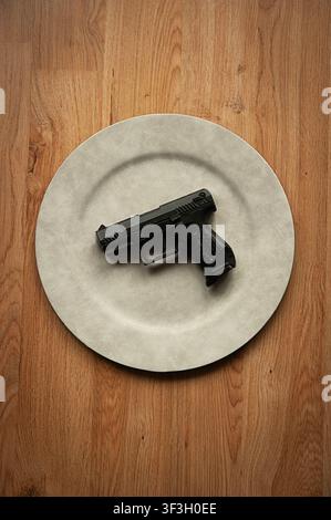 Black handgun placed on an empty white plate on a wooden floor, a conceptual still life about violence, weapons and consumption Stock Photo