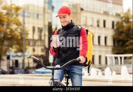 Delivery Guy With Phone Sitting On Bicycle Using App Outdoor Stock Photo