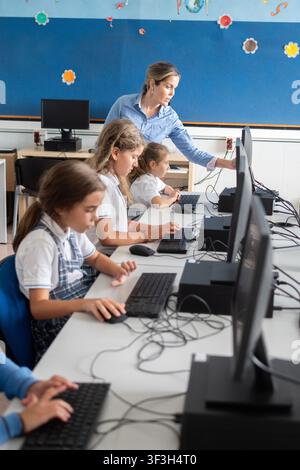 Teacher monitoring elementary school students while they work on desktop computers in a modern computer classroom. Digital learning and back to school Stock Photo