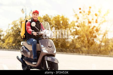 Courier Delivering Food In Yellow Backpack Riding Motorbike Outside Stock Photo
