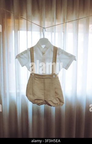Elegant vintage-style baby boy outfit with linen suspenders hanging in natural light. Stock Photo
