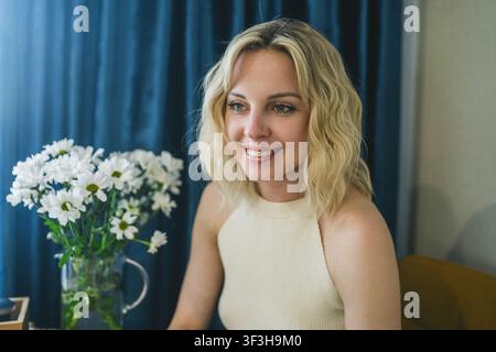 Smiling blonde woman in cream top with white daisies and blue curtain Stock Photo