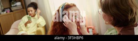 Header of young Caucasian woman receiving facial treatment from middle aged Caucasian beautician while friend using smartphone nearby, promoting skincare service and beauty salon marketing Stock Photo