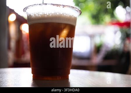 Backlit iced americano coffee with pineapple juice in clear plastic cup, condensation droplets and frothy top on outdoor cafe table Stock Photo