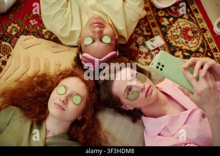 Overhead shot of young adult women relaxing with cucumber slices and eye patches, taking smartphone selfie during home spa session. Ideal for skincare, self care, friendship marketing Stock Photo