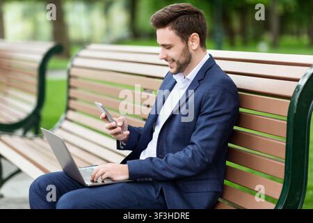 Successful businessman working online using laptop computer and smartphone on bench in park Stock Photo