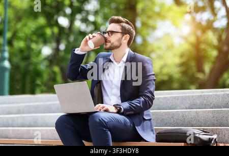 Corporate employee drinking coffee while using laptop on stairs at park Stock Photo
