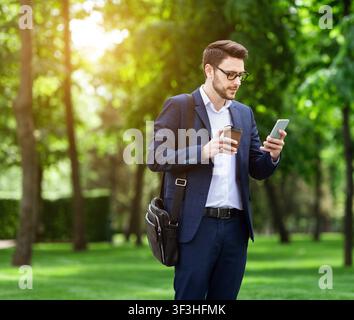 Young business manager with takeout coffee browsing net on cellphone at city park Stock Photo