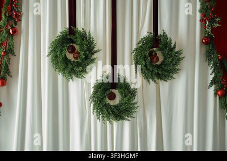 holiday photo studio backdrop featuring a draped white fabric background flanked by red curtains, pine garlands with red ornaments, and three small Ch Stock Photo