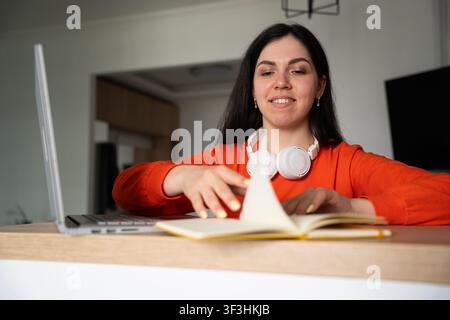 Young woman using laptop and notes in home office environment. Online learning tutoring and coffee break productivity. Stock Photo