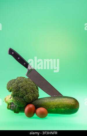 Colorful vertical studio still life with a chef knife, broccoli, zucchini and brown eggs arranged on a mint green background with copy space. The red Stock Photo