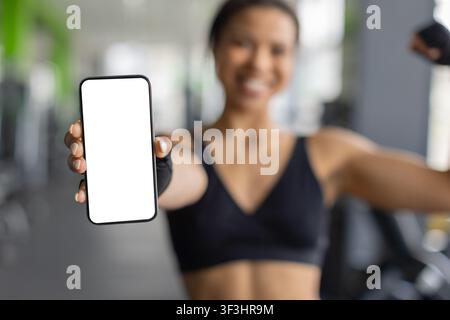 Smiling young Black woman flexing her arm and showing blank smartphone screen Stock Photo