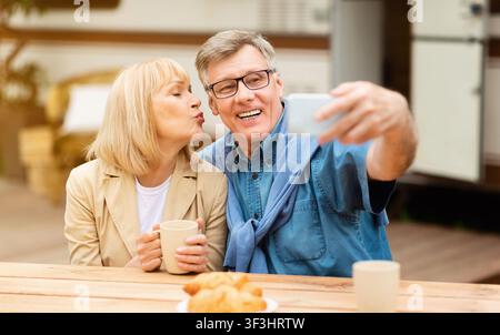 Mature woman kissing her husband while he taking selfie near motorhome outdoor Stock Photo