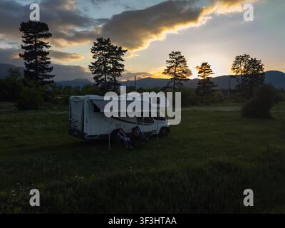 Couple enjoying the outdoor lifestyle, sitting under an awning next to their motorhome in a grassy field, watching the beautiful sunset behind distant Stock Photo